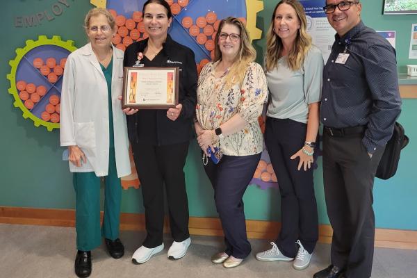 Ivonne Rodriguez receiving award standing with Dr. Freedman, Ashley Grant, Valerie Honeycutt and Cesar Salazar in front of a toy gear on the wall