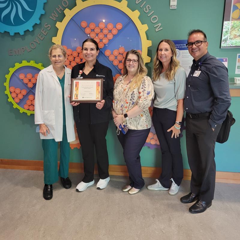 Ivonne Rodriguez receiving award standing with Dr. Freedman, Ashley Grant, Valerie Honeycutt and Cesar Salazar in front of a toy gear on the wall