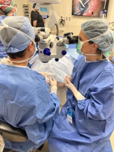 Eye surgery photo with eye surgeon Lloyd Williams at the head of the patient teaching a visiting doctor sitting to the side how to do surgery. 